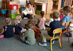Dzieci siedzą na dywanie i słuchają książki czytanej przez mamę dziecka.