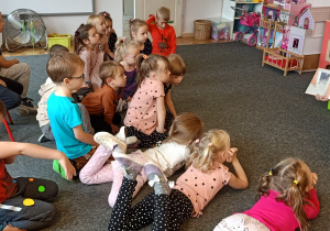 Dzieci siedzą na dywanie i słuchają książki czytanej przez mamę dziecka.