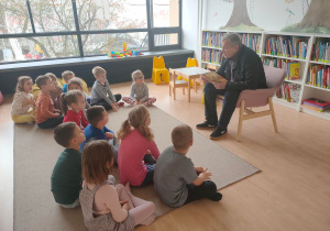 Dzieci siedzą na dywanie w bibliotece i słuchają książki czytanej przez aktora.