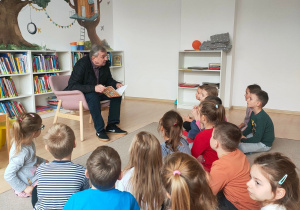 Dzieci siedzą na dywanie w bibliotece i słuchają książki czytanej przez aktora.