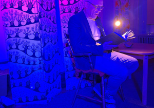 Aktor czytający historię z książki o Kubusiu Puchatku siedzi na eleganckim krześle przy zapalonych lampkach.