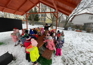 Dzieci zebrane wraz z nauczycielką pod wiatą w punkcie dydaktycznym w ogrodzie przedszkolnym.