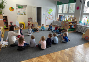 Grupa dzieci zebrana na dywanie w sali, słucha prowadzącej zajęcia.